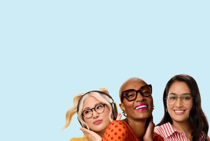 Group portrait of three stylish women wearing eyeglasses against a solid light blue background, including a blonde woman with headphones in round spotted frames, a smiling woman with short hair in thick square tortoiseshell frames, and a smiling woman in a striped shirt wearing thin metal aviator frames.