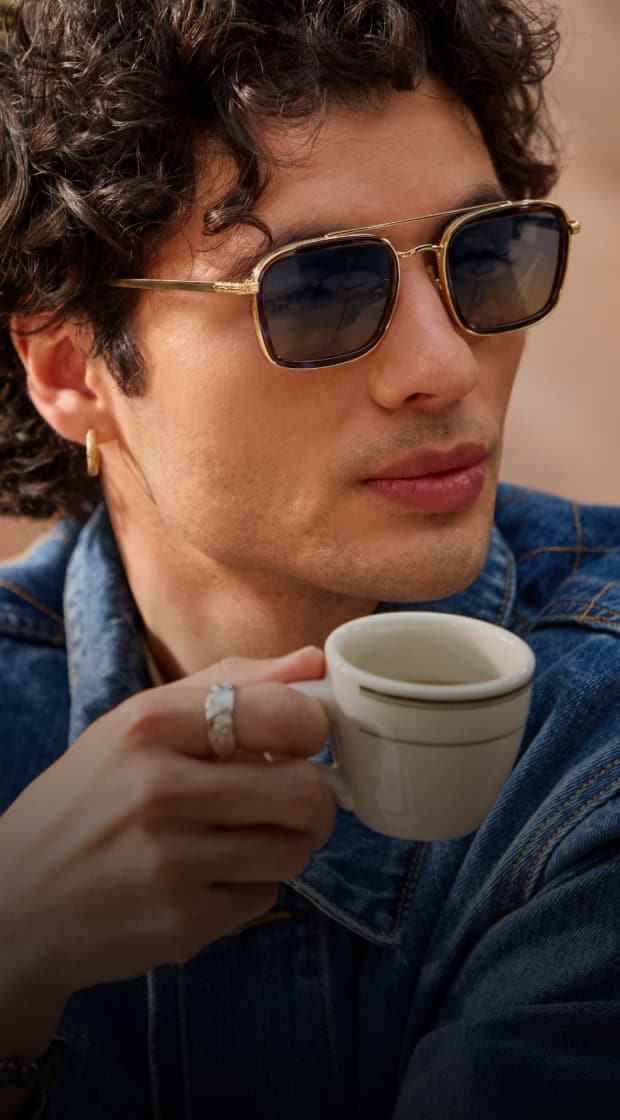 A man in gold square aviator Zenni sunglasses. He has mid-length curly brown hair and is sipping an espresso. Teal overlay design with leaf shadows.