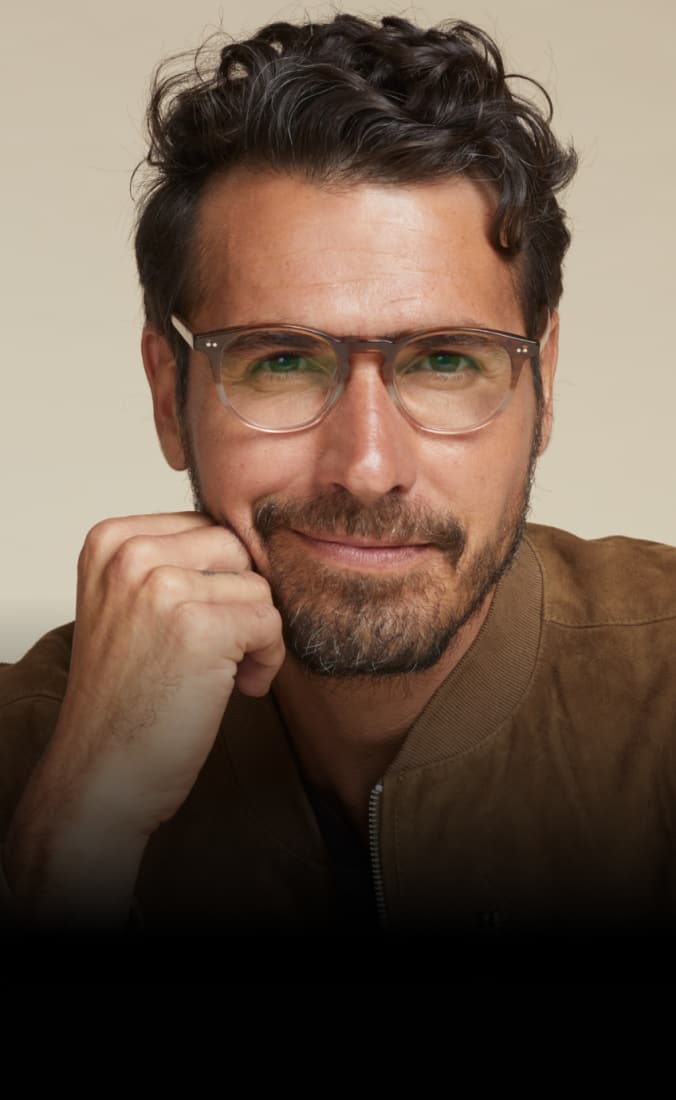 A man with short, curly dark hair and a trimmed beard smiles subtly, resting his chin on his hand. He is wearing brown two-tone glasses and a tan suede jacket over a black shirt.