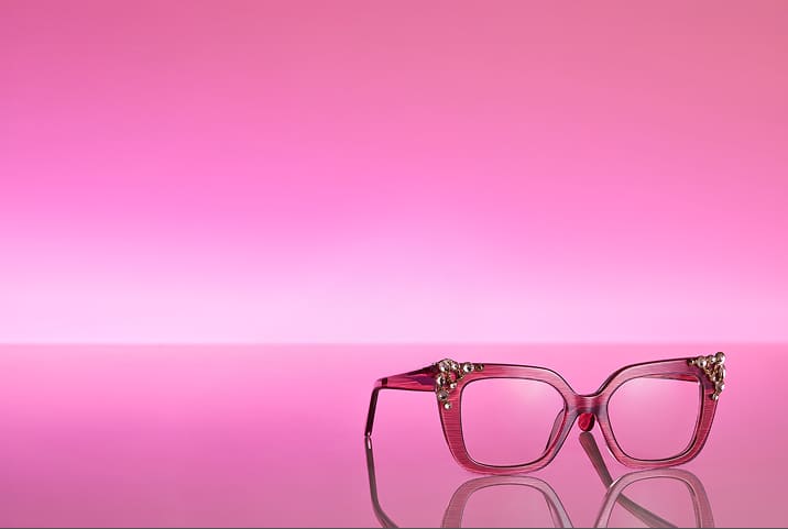 Red eyeglasses with metallic embellishments against a pink gradient background.
