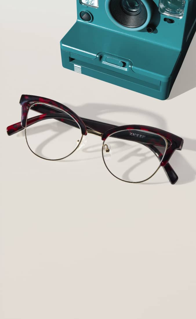 Red and black tortoiseshell eyeglasses with gold accents style no. 1916225 on a light background. A teal instant camera is positioned in the top right corner.