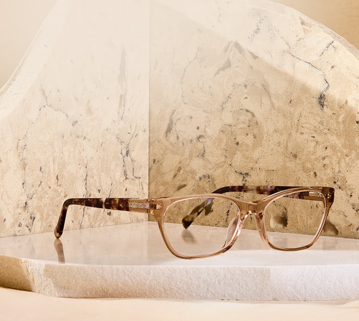 A pair of translucent champagne rectangle glasses with a subtle pink tint, featuring glittery rose-gold detailing on the temples and tortoiseshell arms, resting on a white stone pedestal against a textured beige background.
