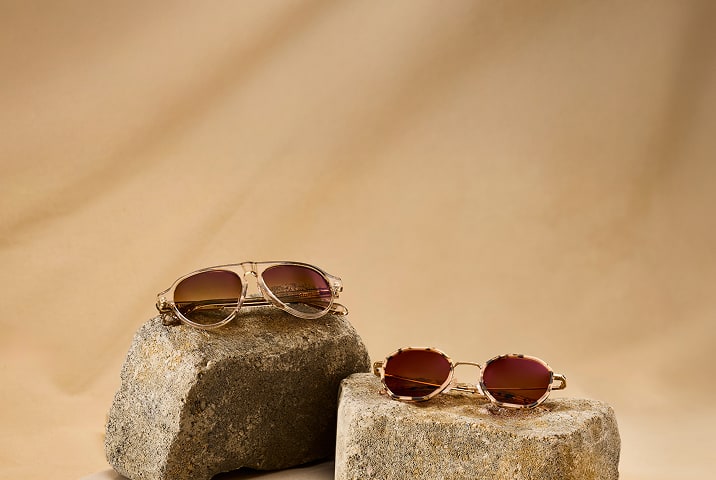 Light brown aviator sunglasses with brown gradient lenses and geometric tortoiseshell Zenni glasses with brown lenses, on a beige stone background.