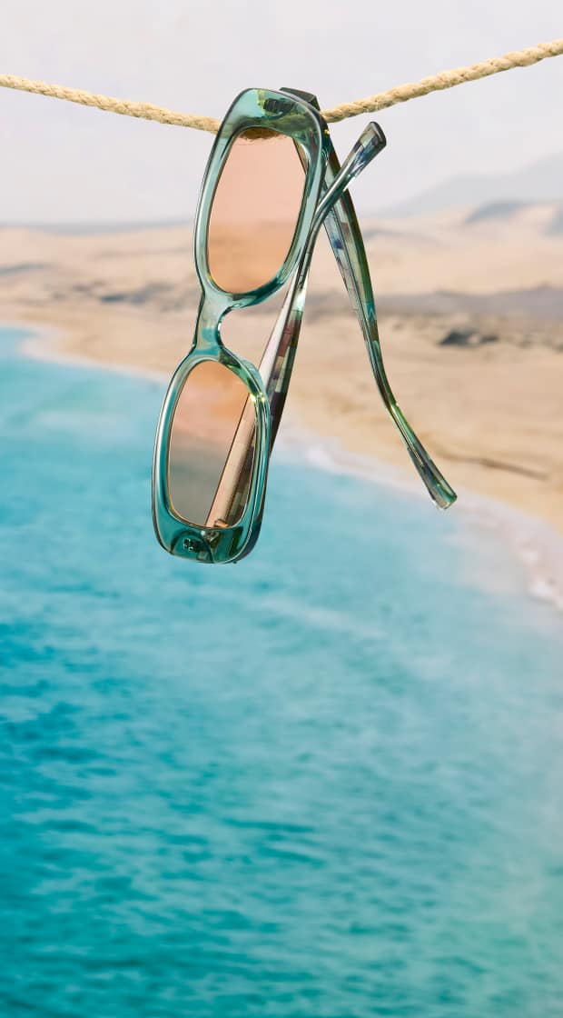 Rectangle transparent Zenni sunglasses with pale amber lenses and mosaic pattern temples, hanging on a rope in front of a coastal landscape with sand dunes.