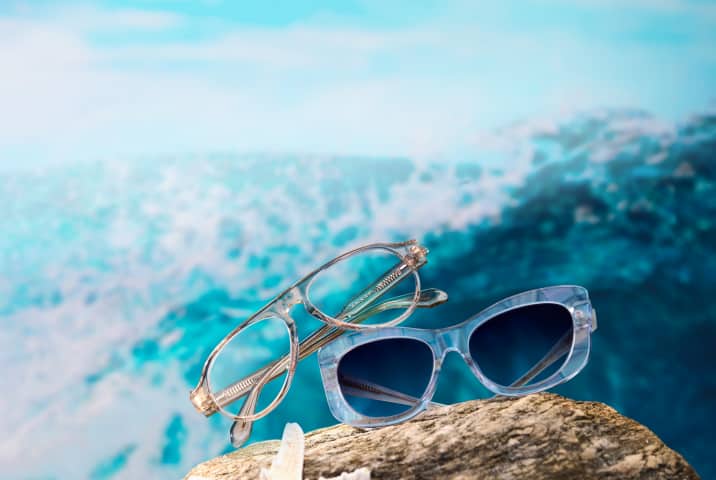 Gafas de aviador translúcidas y gafas de sol cuadradas de color azul claro, posadas sobre una roca junto a una estrella de mar blanca, con un fondo borroso de océano turquesa.