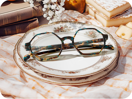 Geometric brown and blue tortoiseshell glasses resting on an antique china plate, on a pale pink picnic blanket with vintage books, brie cheese, and white flowers nearby.