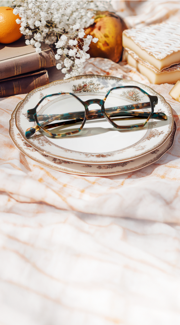 Geometric brown and blue tortoiseshell glasses resting on an antique china plate, on a pale pink picnic blanket with vintage books, brie cheese, and white flowers nearby.