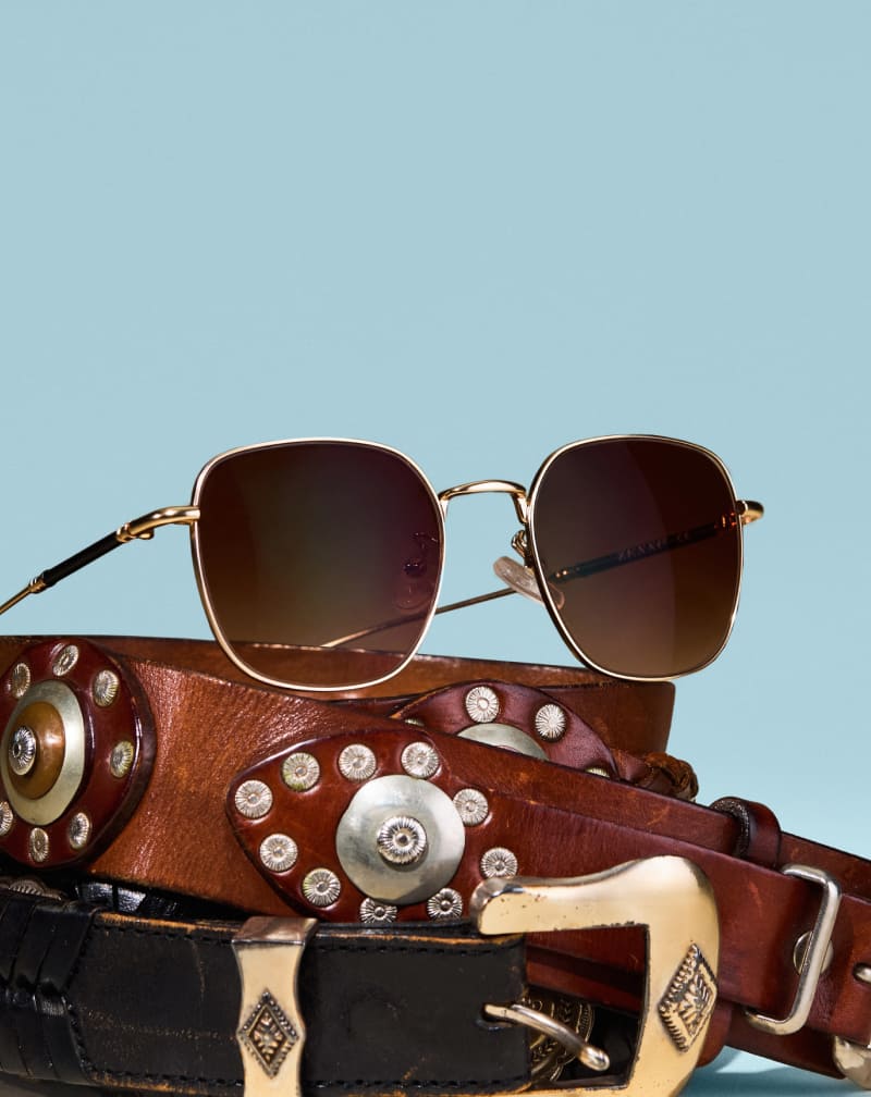 Gold wireframe sunglasses with brown gradient lenses resting on studded leather western belts, set against a light blue background.