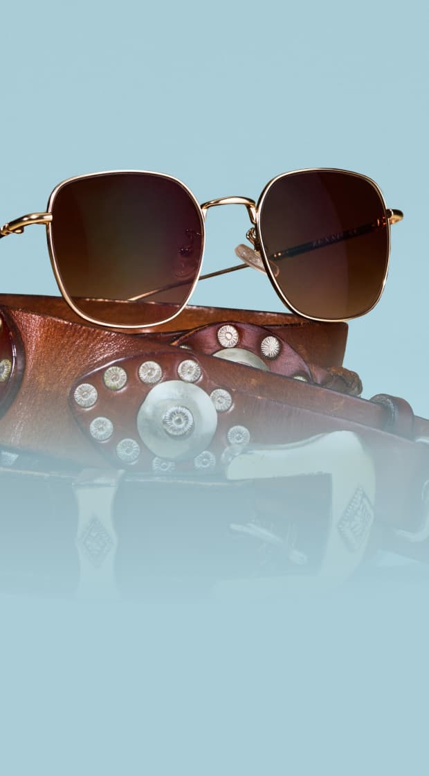 Gold wireframe sunglasses with brown gradient lenses resting atop a embellished brown leather western belt against a light blue background.