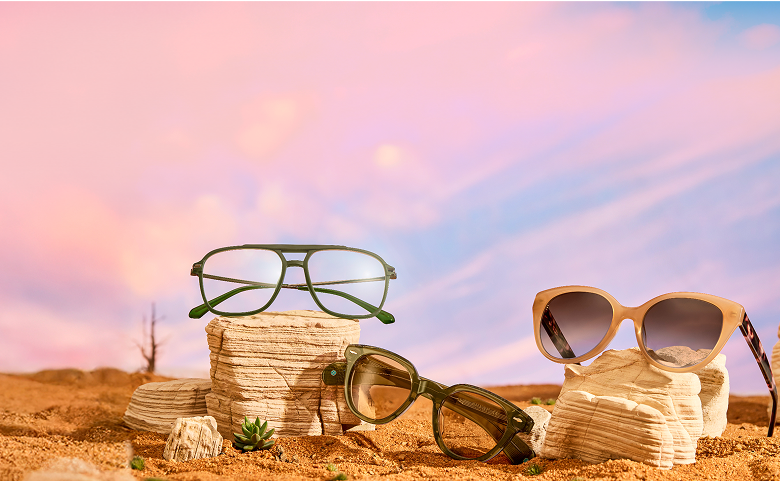 Three pairs of stylish sunglasses are displayed on layered rock formations in a desert-like setting, with a dramatic pink and blue sky in the background.