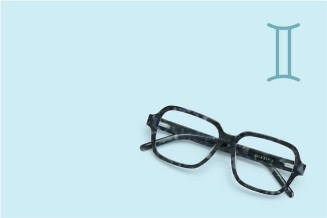 A pair of dark patterned acetate glasses on a light blue background with gemini sign in the top right corner.