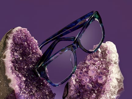 Multicolor purple square glasses resting on a big amethyst geode, on a purple background.