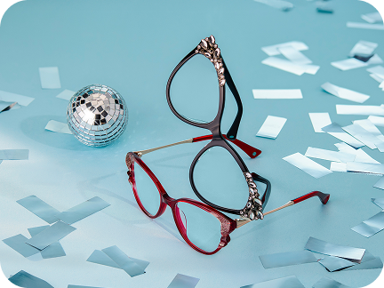 Two pairs of glasses, one black and one red, both with rhinestones on the corners, are stacked on a light blue surface next to a small disco ball, scattered with silver confetti.