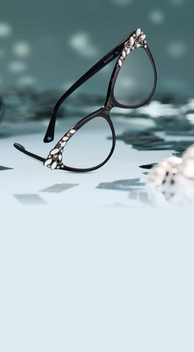 A close-up of a pair of black cat-eye glasses, embellished with clear jewels on the corners, resting on a shiny surface surrounded by silver confetti and small disco balls.