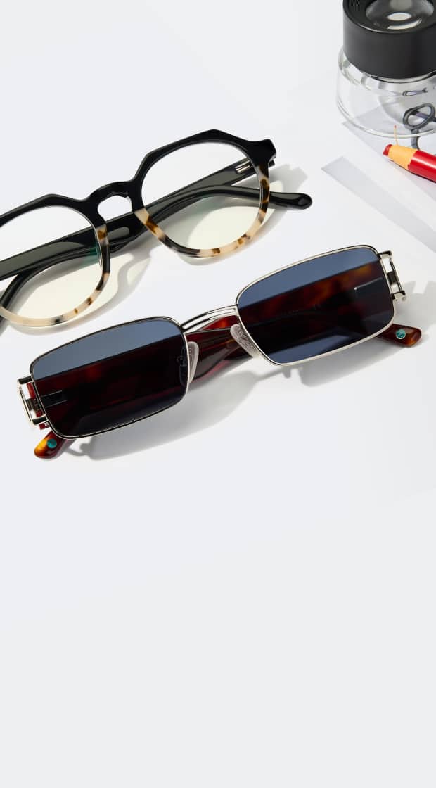 Geometric black and tortoiseshell glasses paired with slim rectangular silver sunglasses and desk accessories on a white surface.