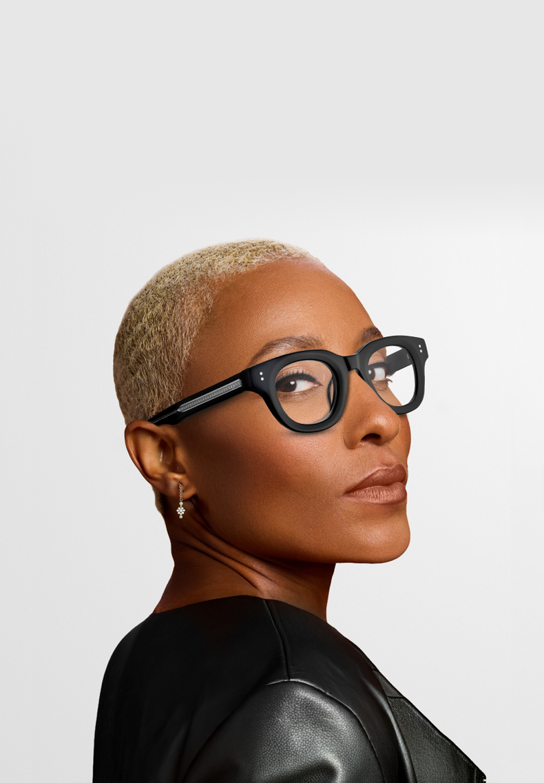 A profile portrait of a woman with short blonde hair wearing bold, thick-rimmed black rectangular eyeglasses and a black leather jacket.