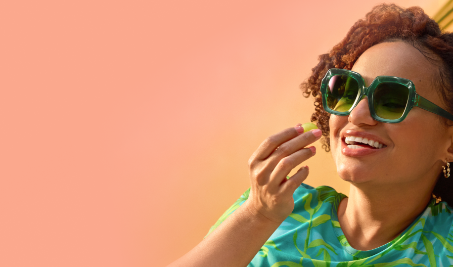 A smiling woman with curly hair and a teal flower accessory wearing oversized green geometric sunglasses, set against a warm orange and pink gradient summer background.