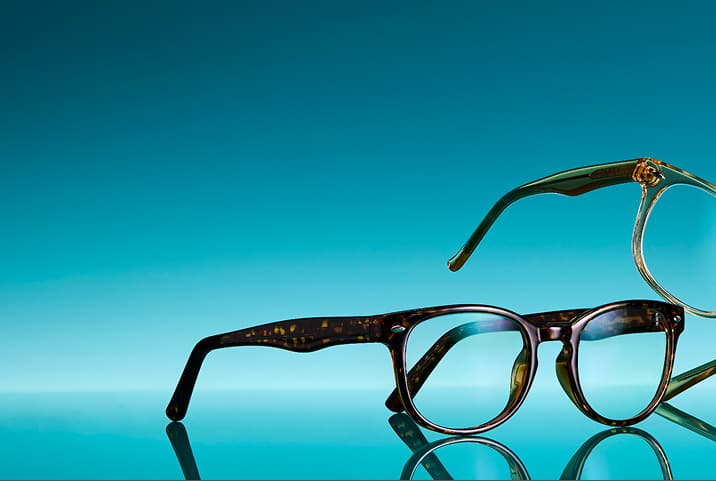 Two pairs of Zenni eyeglasses in trending frame styles and colors on a glossy aqua surface.