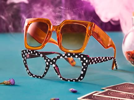 Orange oversized square sunglasses stacked on a pair of black and white polka dot square glasses, on a light teal table, surrounded by dried flowers and tarot cards. Eerie purple smoke floats in the background.