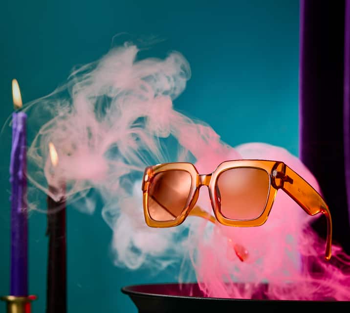 Orange oversized square sunglasses floating above a smoking cauldron, with black and purple taper candles nearby.