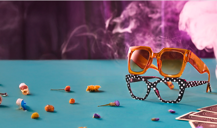 Orange oversized square sunglasses stacked on a pair of black and white polka dot square glasses, on a light teal table, surrounded by dried flowers, apothecary jars, and tarot cards. Eerie purple smoke floats in the background.
