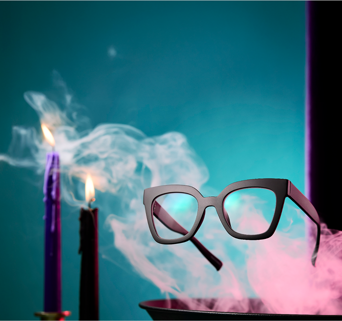 Black cat-eye oversized glasses floating above a smoking cauldron, with black and purple taper candles nearby.