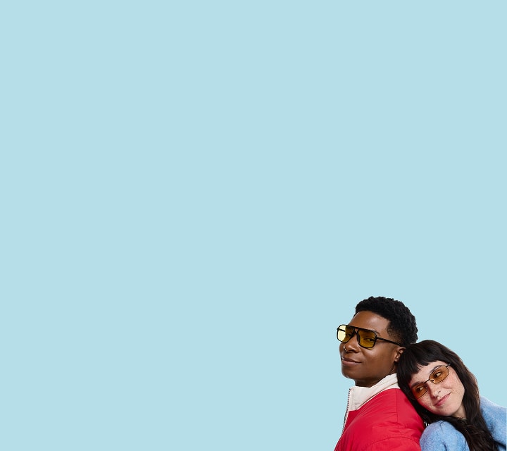 A man wearing bold black frames with yellow-tinted lenses and a red jacket smiles while a woman with bangs, wearing rectangular frames with amber-tinted lenses and a blue sweater, leans her head on his shoulder against a light blue background.