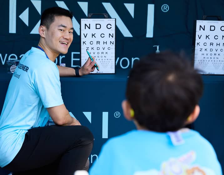 Evaluador visual masculino sonriente sosteniendo un bolígrafo verde y señalando una tabla optométrica con letras blancas mientras mira hacia un niño pequeño sentado en primer plano, con un banner de la marca 'ZENNI' en el fondo.