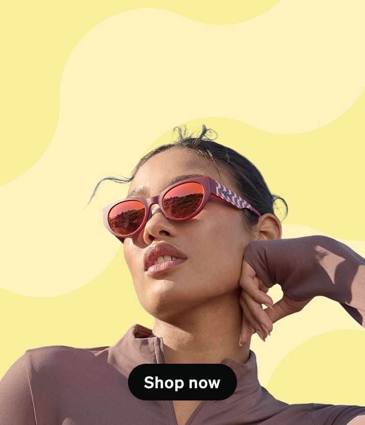 Vista desde un ángulo bajo de una mujer con el cabello recogido en un chongo mirando hacia arriba mientras usa lentes de sol estilo ojo de gato con montura roja oscura y cristales reflectantes naranjas, sobre un fondo amarillo pálido con olas abstractas.