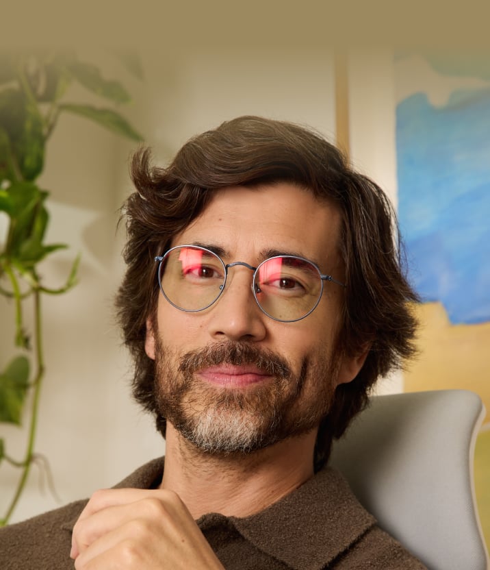 Portrait d'un homme souriant aux cheveux et à la barbe marron portant des lunettes rondes en fil métallique bleu dans une pièce lumineuse avec des plantes d'intérieur.
