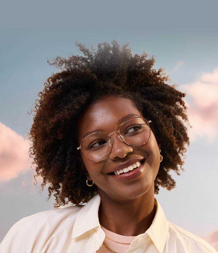Jeune femme souriante aux cheveux naturellement bouclés portant des lunettes géométriques en fil métallique doré, regardant vers le haut sur fond de ciel au coucher du soleil.
