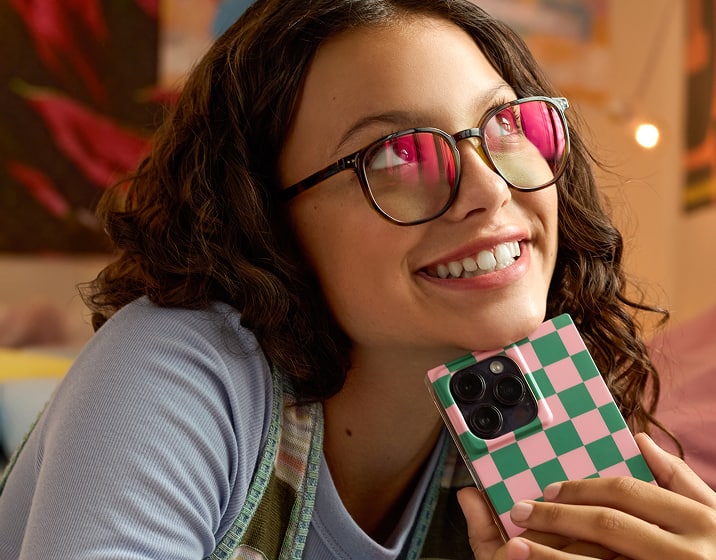 Jeune femme souriante aux cheveux foncés et bouclés portant des lunettes rondes en écaille, posant son menton sur un smartphone à coque damier vert et rose tout en regardant vers le haut.