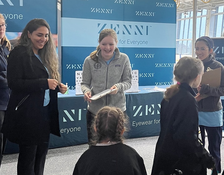 A group of Zenni volunteers holding clipboards smiling at children seated for their vision screenings.