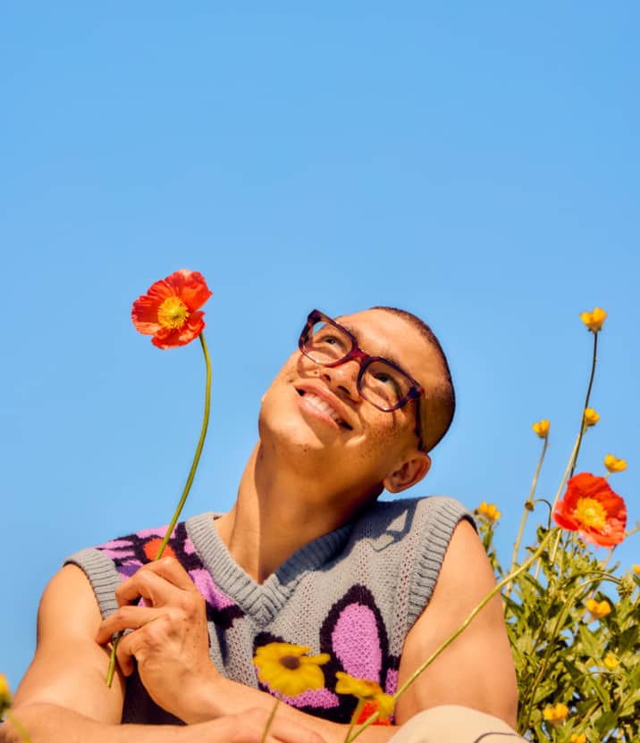 A person wears EcoBloomz eyeglasses while holding red poppies in a wildflower field, with text noting that proceeds support 1% for the Planet.