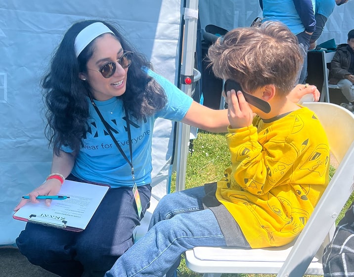 A Zenni volunteer kneels beside a young boy at an outdoor vision screening event.