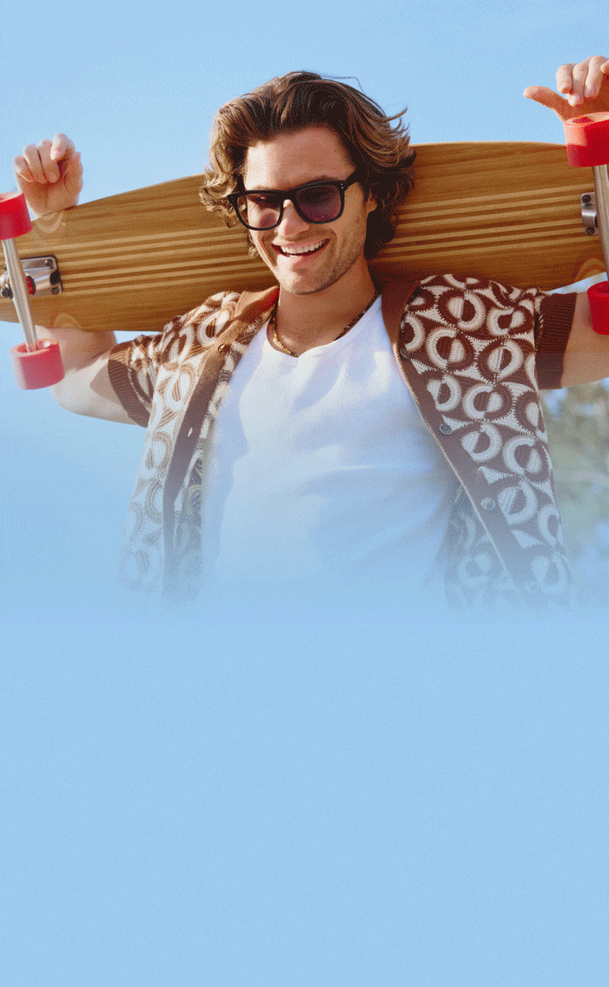 Chase Stokes in Zenni Jetsetter sunglasses, smiling outdoors in a white and brown geometric shirt against a blue sky with a wooden skateboard over his shoulders.