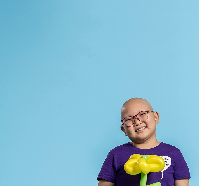 A smiling child wearing round, pink glasses and a purple shirt with a white heart graphic. They are holding a yellow and green balloon shaped like a flower. The background is a solid light blue, creating a cheerful and uplifting mood.