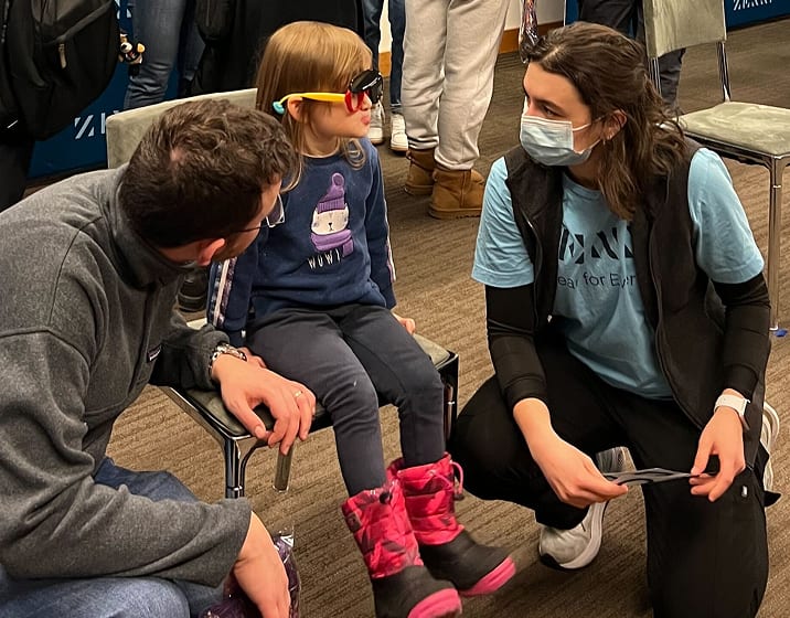 A young child wearing red and yellow glasses sits on a chair while a man and a woman in a medical mask and Zenni shirt kneel beside them to assist during an eye care event.