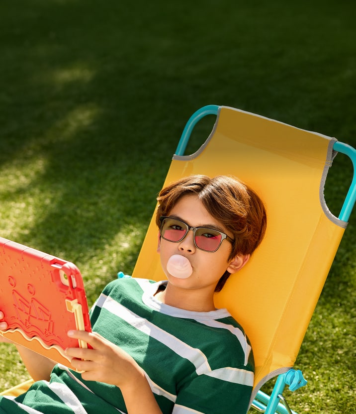 A boy with brown hair wearing black rectangle glasses with gray EyeQLenz lenses that have a slight pink sheen. He’s sitting on a yellow lawn chair, reading on a tablet, and blowing a bubble with pink bubblegum.