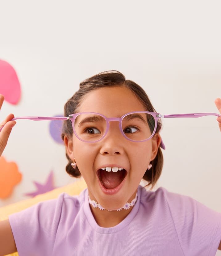 A girl with dark brown hair pulls on the temple arms of her pink flexible hinge square glasses. She is smiling in amazement.