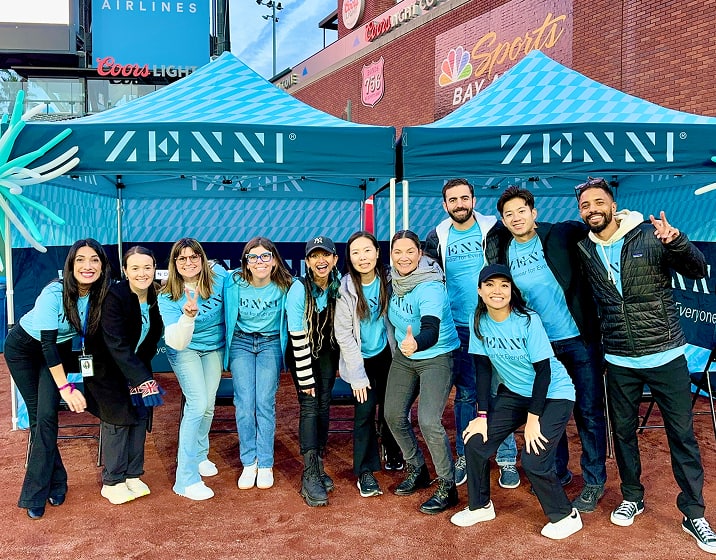 A group of eleven smiling people, mostly wearing light blue shirts with a Zenni logo, posing for a photo in front of two dark teal tents with a light blue and white pattern.