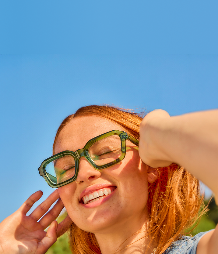 Close-up of a smiling woman in oversized Zenni EcoBloomz green glasses under bright sunlight.