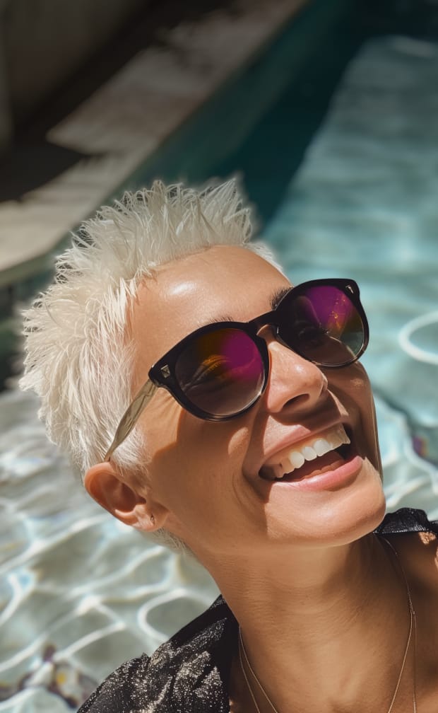 Close-up of a person with short white-blonde hair and stylish frames, facing the sun by a swimming pool, highlighting sun protection.