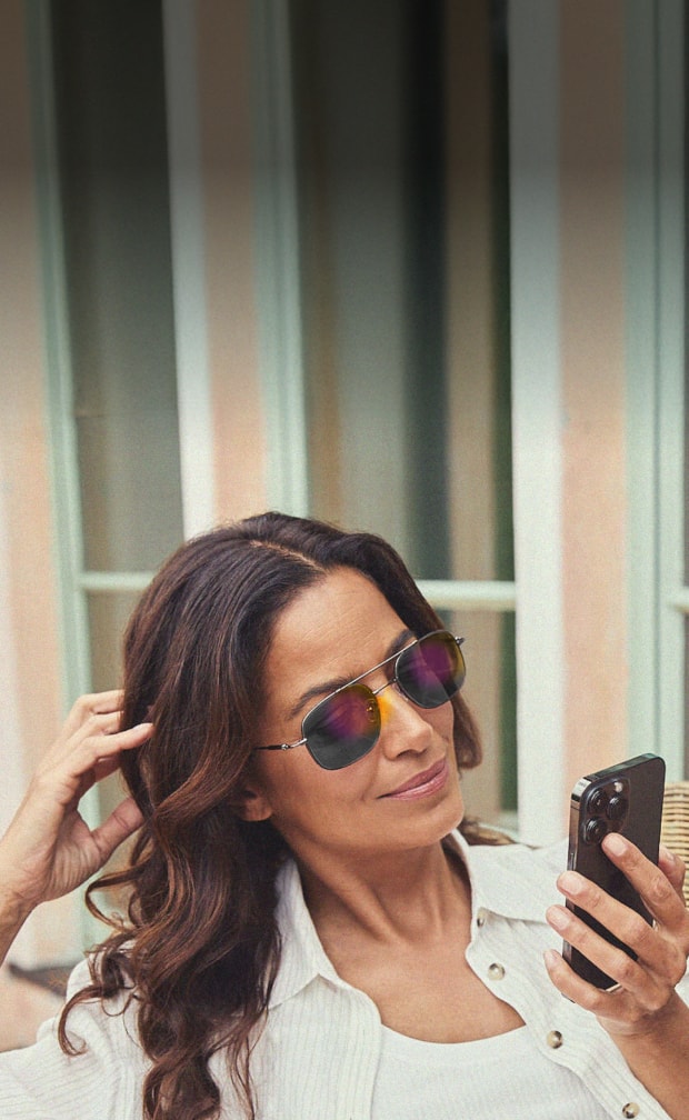 A woman with dark wavy hair focused on her phone and wearing silver aviator glasses, showcasing their blue light filtering function.