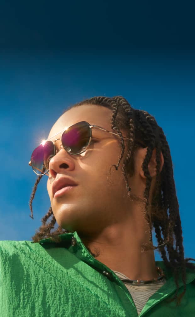 A fashion-forward individual with braids wearing gold aviator EyeQLenz frames from Zenni, gazing pensively into the distance under the sunlight.
