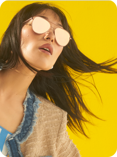 Close-up of a young woman with long brown hair wearing Zenni mirrored Premium Aviator Sunglasses against a bright yellow background.