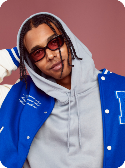 Young man with braids wearing Zenni Rectangle Glasses with FL-41 lenses, a gray hoodie, and blue & white jacket, posing against a rose-colored background.