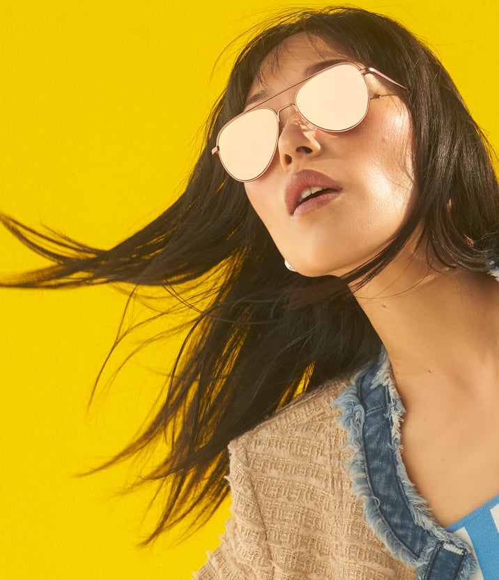 Close-up of a young woman with long brown hair wearing Zenni mirrored Premium Aviator Sunglasses against a bright yellow background.
