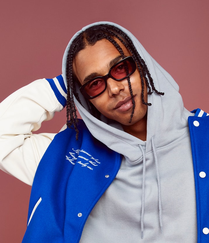 Young man with braids wearing Zenni Rectangle Glasses with FL-41 lenses, a gray hoodie, and blue & white jacket, posing against a rose-colored background.