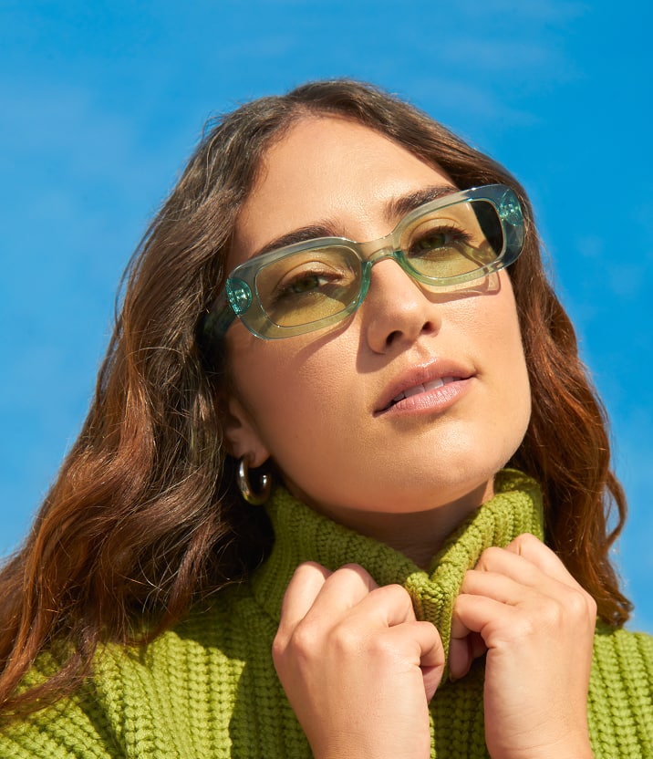 A young woman with long brown hair wearing translucent green Zenni rectangle glasses and a green turtleneck against a bright blue background.